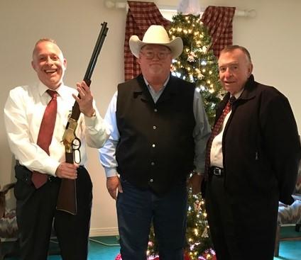 Two men in suits, one holding a shotgun pointed at the ceiling, standing on either side of Sheriff Singleton